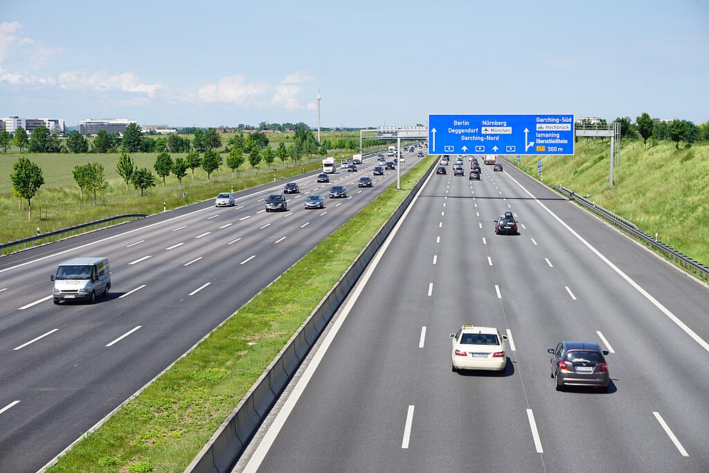 A photo of a German autobahn (A9) near Munich showing clear lane discipline with multiple lanes in both directions. The highway features crisp white lane markings, minimal traffic, and blue directional signs indicating routes to Berlin, Nürnberg, and other destinations. The organized traffic flow demonstrates how structured constraints enable efficient movement.